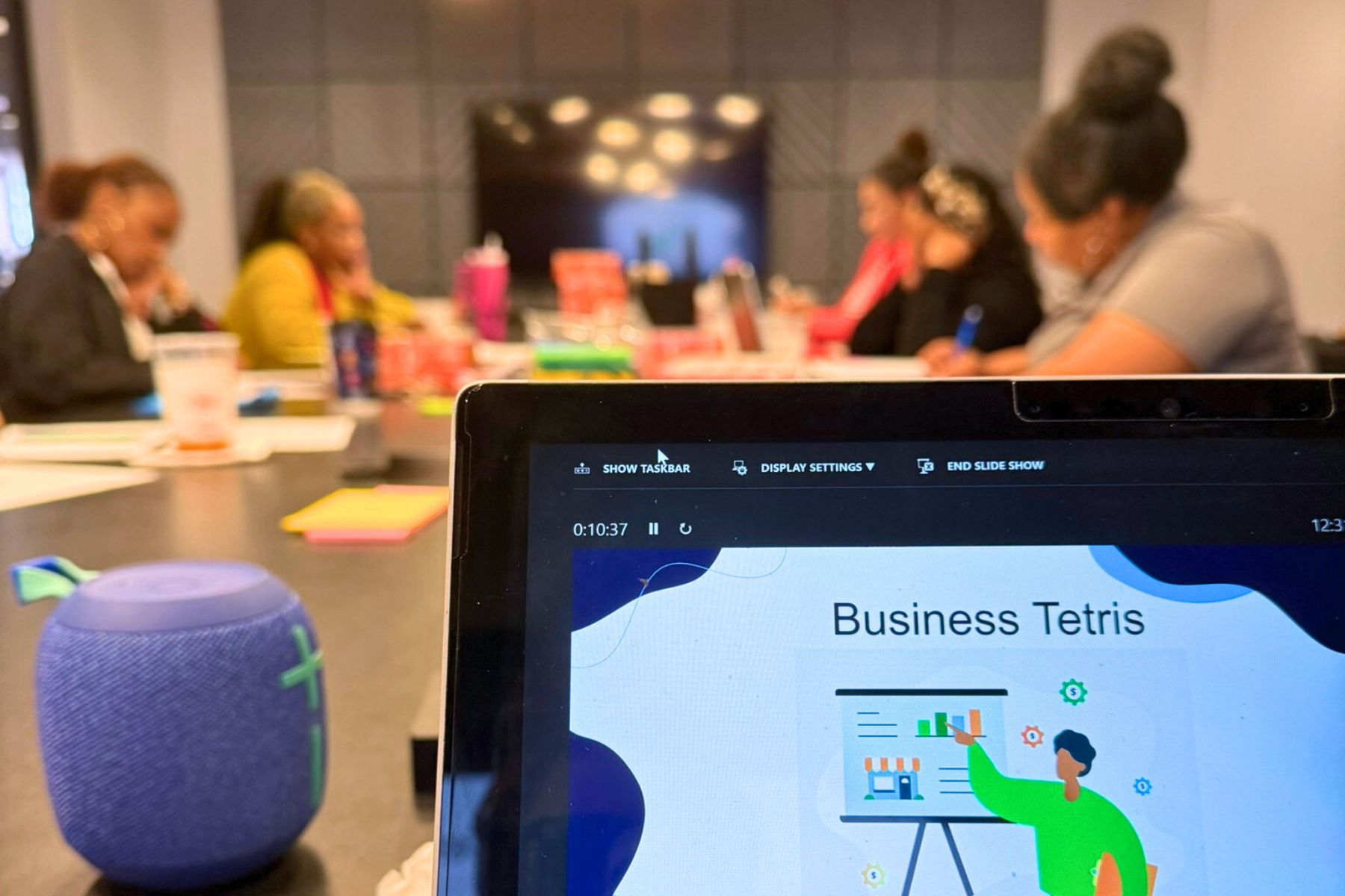 “Business Tetris” slide on a laptop, educators working in the background (blurred).